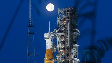 Naktį ant NASA kosminio paleidimo sistemos raketos viršaus pasirodo didelė ir labai ryški pilnatis. Oranžinės ir baltos spalvos raketa stovi šalia plieninės atraminės konstrukcijos, vadinamos mobiliuoju paleidimo įrenginiu. NASA Naktį ant NASA kosminio paleidimo sistemos raketos viršaus pasirodo didelė ir labai ryški pilnatis. Oranžinės ir baltos spalvos raketa stovi šalia plieninės atraminės konstrukcijos, vadinamos mobiliuoju paleidimo įrenginiu. NASA